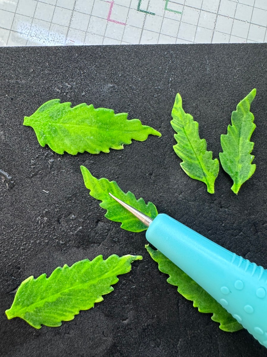 Close-up of green serrated paper leaves being shaped with a Leaf Veining tool on a crafting workspace.