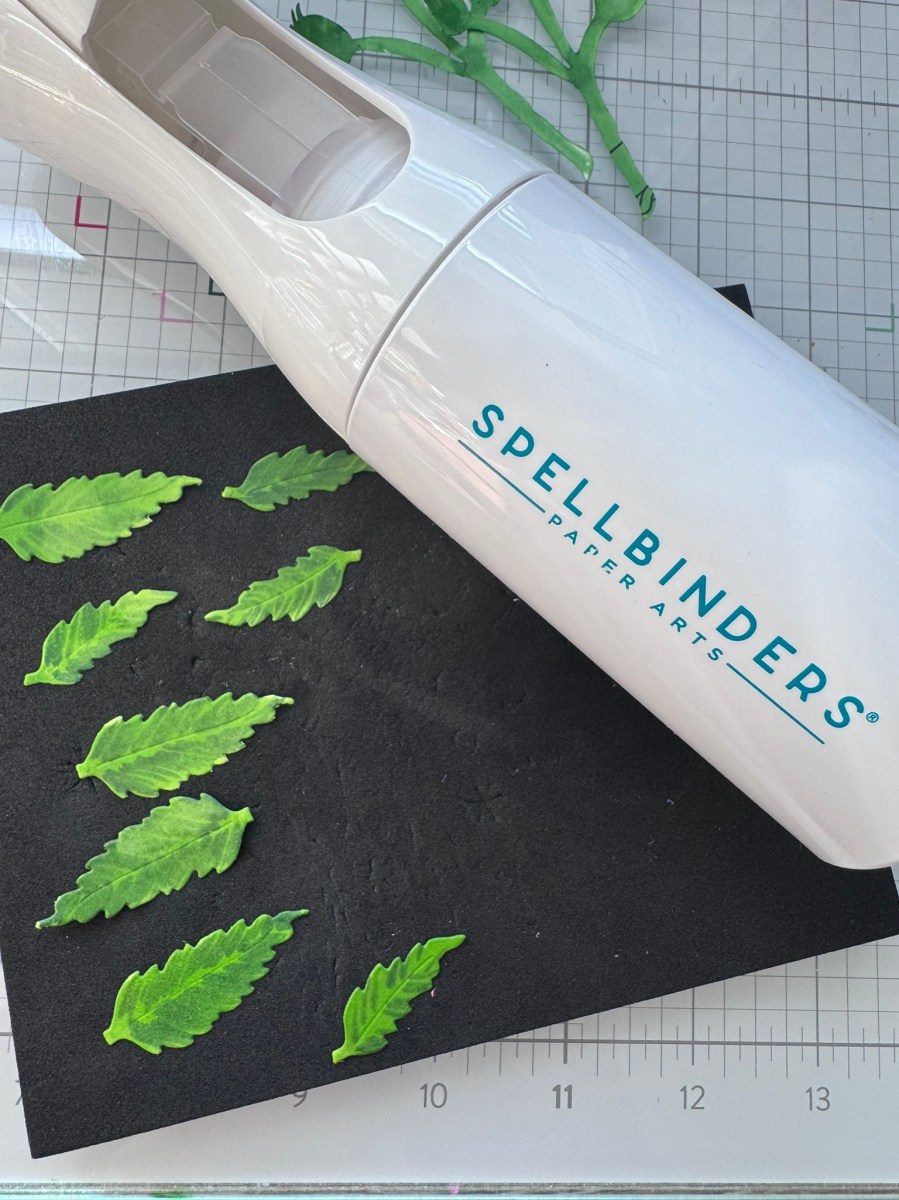 Spellbinders Paper Arts misting bottle resting on a foam mat beside green die-cut leaves for a paper crafting project.