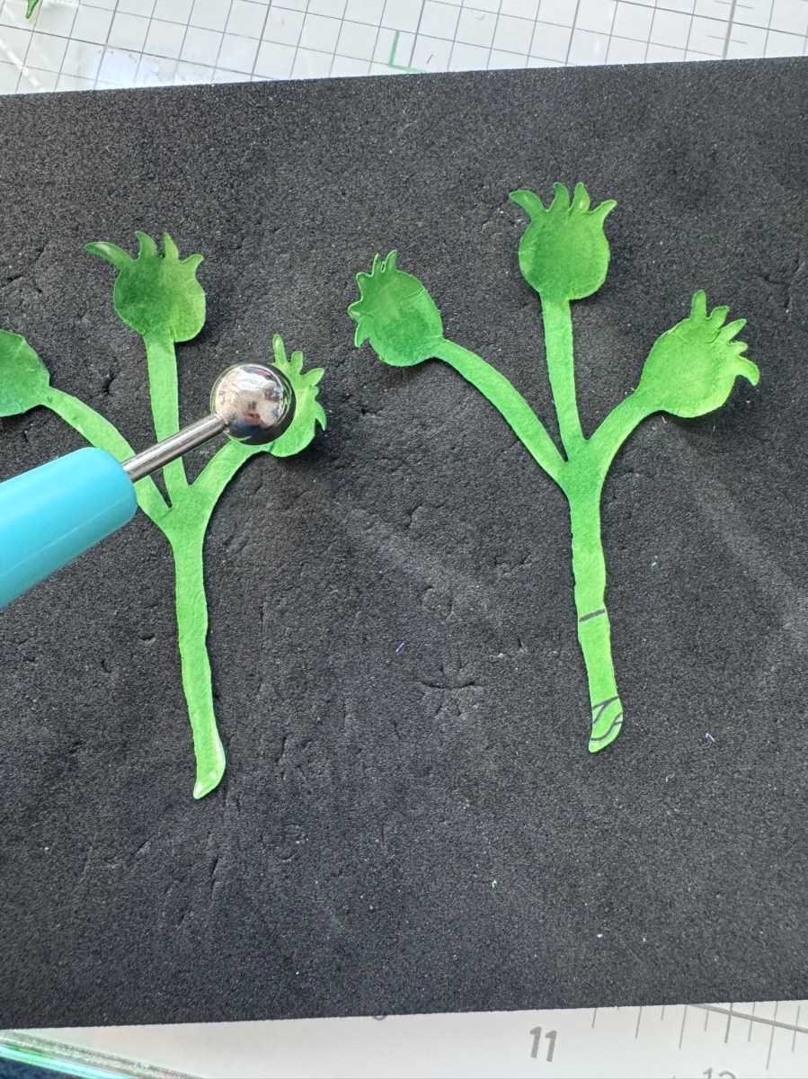 Paper crafting process showing green die-cut stems and buds being shaped with a ball stylus on a foam mat.